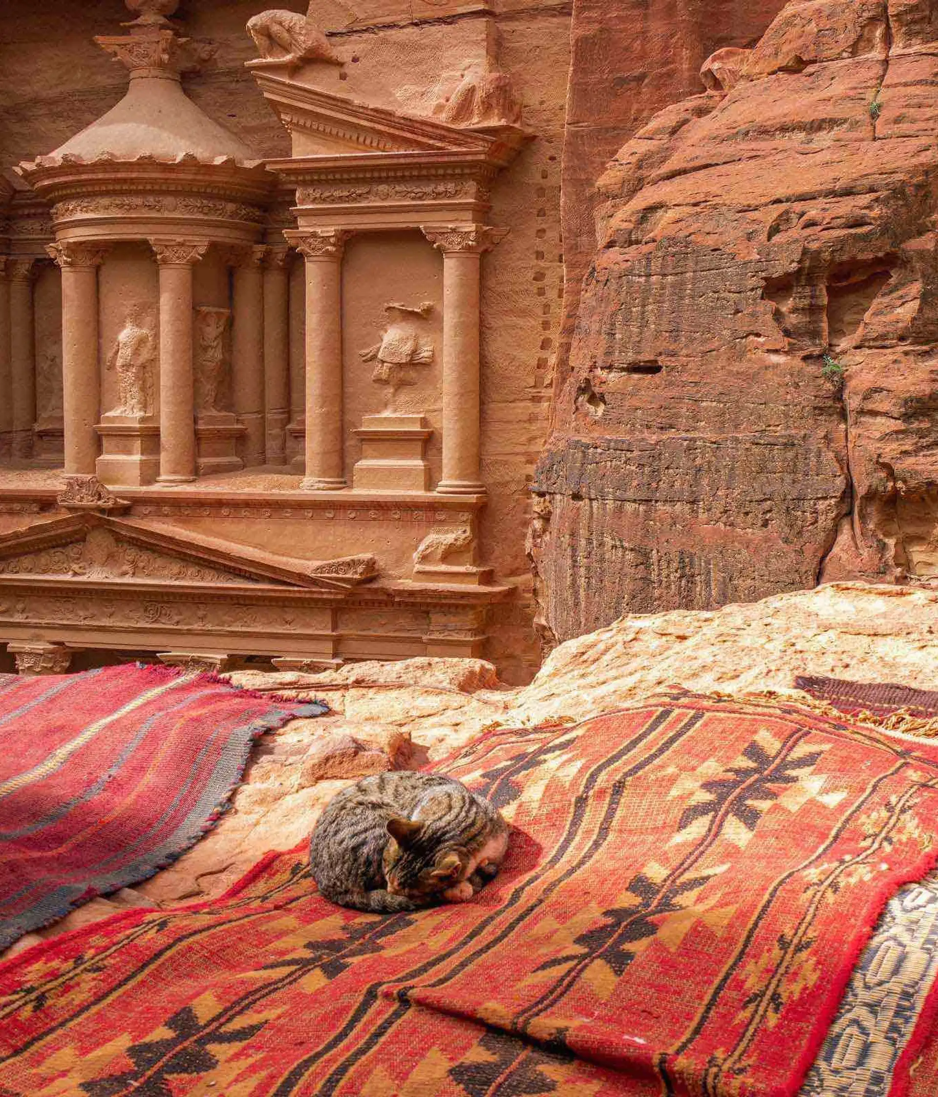 Façade monumentale sculptée dans la roche de grès rose du temple Al-Khazneh (Le Trésor) à Pétra, vue depuis un promontoire avec des tapis traditionnels bédouins au premier plan.