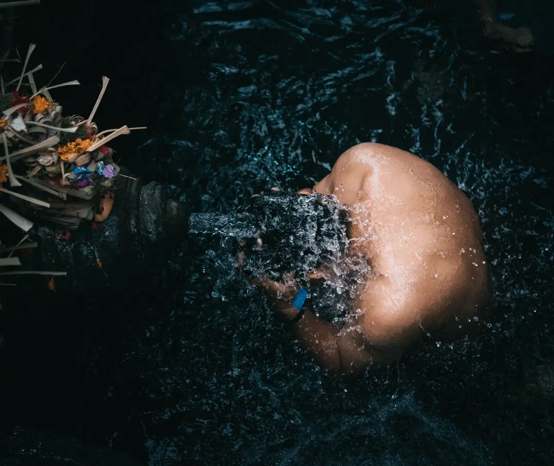 Voyage en Asie – Vue de haut d'une personne se recueillant sous un jet d'eau sacrée d'un bassin en pierre orné d'offrandes à Bali.