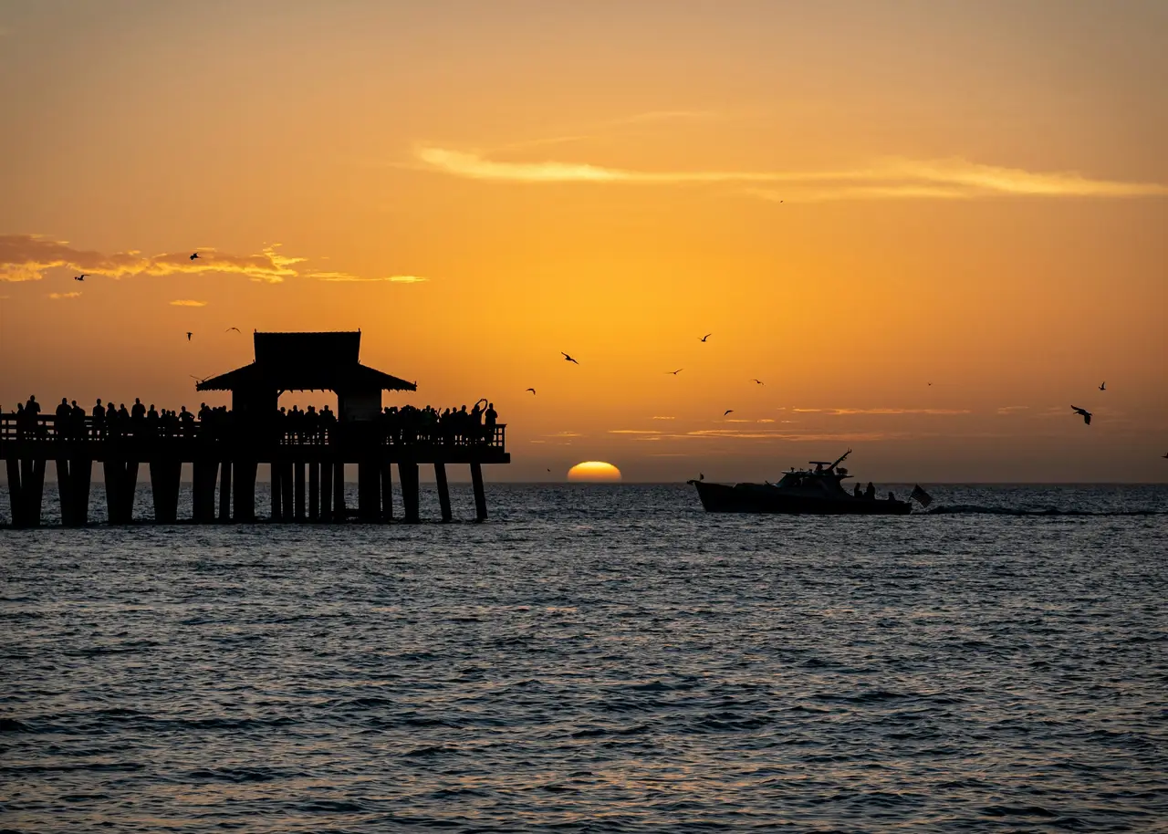 Silhouette de la jetée historique de Naples Pier et d'un bateau sur la mer au coucher du soleil sous un ciel doré et orangé
