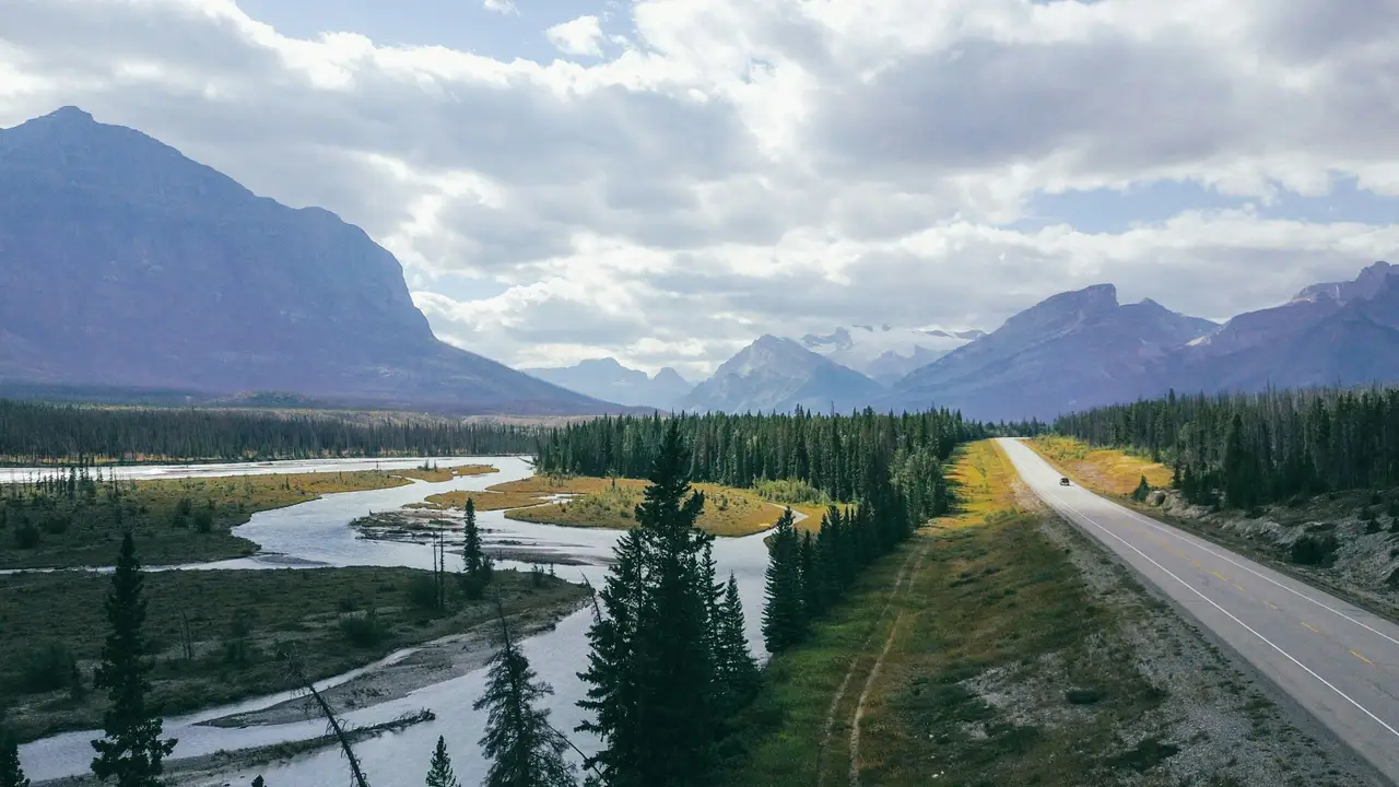 Route asphaltée longeant une rivière et une forêt de conifères bordées d'imposantes montagnes sous un ciel nuageux en Colombie-Britannique