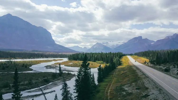 Route asphaltée longeant une rivière et une forêt de conifères bordées d'imposantes montagnes sous un ciel nuageux en Colombie-Britannique