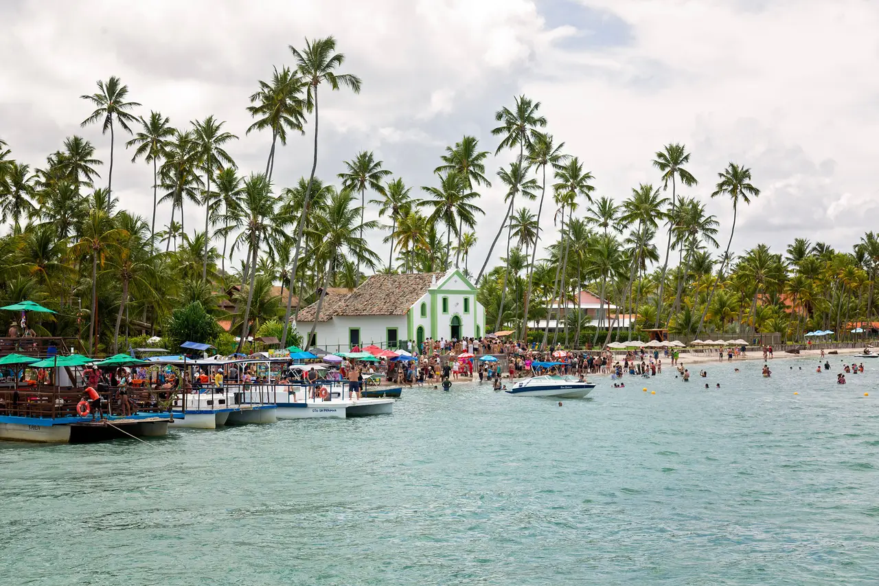Voyage au Brésil – Petite église blanche et verte sur la plage de São Miguel dos Milagres, entourée de grands cocotiers et de bateaux sur l'eau turquoise en Alagoas.