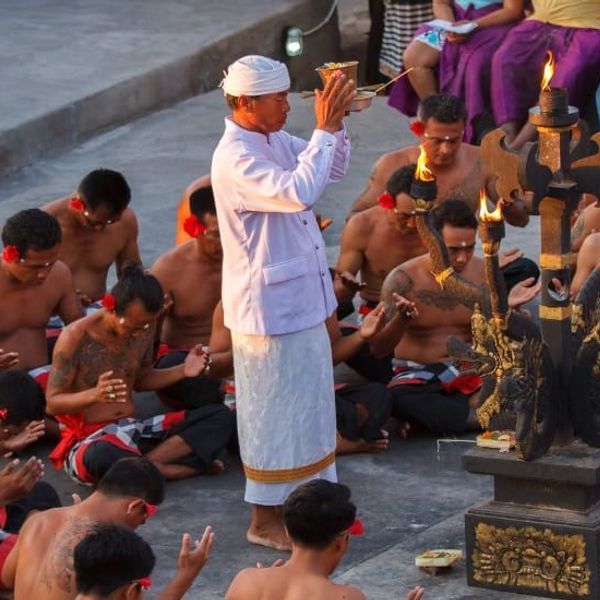 Un prêtre balinais en habit blanc traditionnel dirige une cérémonie de prière en plein air avec des hommes assis en cercle autour d'un autel orné au coucher du soleil à Uluwatu, Bali.