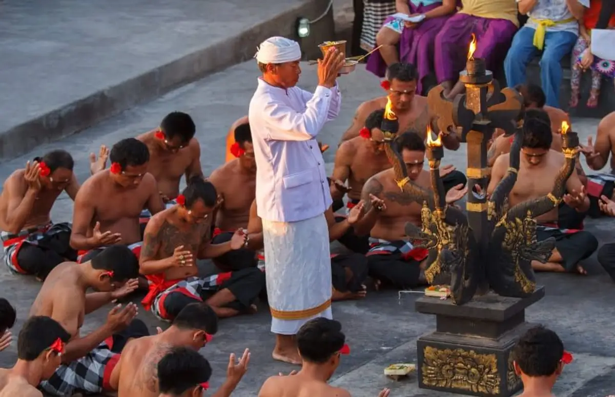 Un prêtre balinais en habit blanc traditionnel dirige une cérémonie de prière en plein air avec des hommes assis en cercle autour d'un autel orné au coucher du soleil à Uluwatu, Bali.