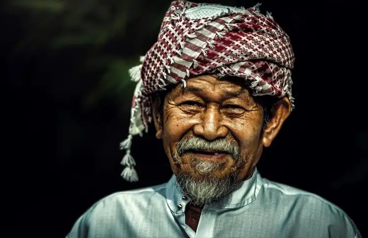 Portrait en gros plan d'un homme âgé souriant avec une moustache, portant un turban traditionnel en tissu à motifs blancs et bordeaux sur fond sombre.