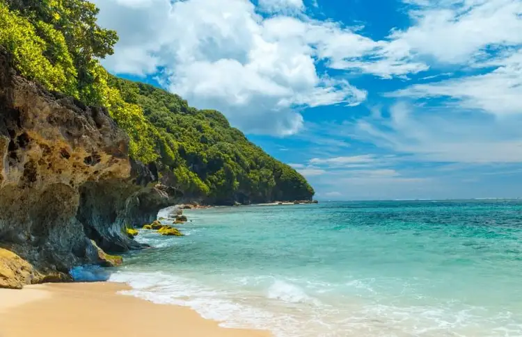 Vue d'une plage de sable doré à Jimbaran, bordée de falaises calcaires accidentées couvertes de végétation luxuriante, avec des vagues d'eau turquoise sous un ciel bleu nuageux à Bali.