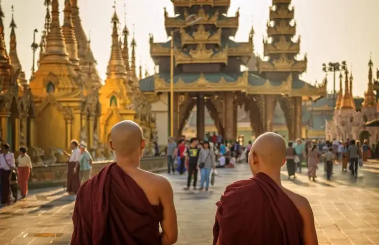 Deux moines bouddhistes en robe traditionnelle bordeaux vus de dos, marchant vers l'entrée d'un complexe de temples bouddhistes aux stupas dorés étincelants à Yangon en Birmanie.