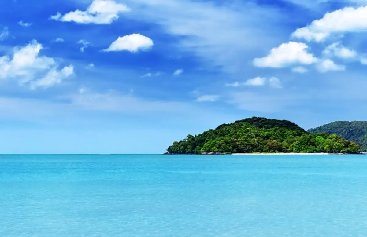 Une colline tropicale couverte d'une végétation verdoyante bordée par une mer turquoise et calme sous un ciel bleu parsemé de nuages blancs à l'île de Langkawi en Malaisie.
