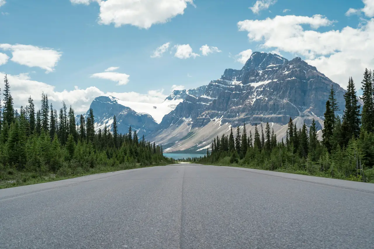 Route asphaltée traversant la forêt boréale en direction des hauts sommets rocheux sous un ciel bleu nuageux près de Jasper