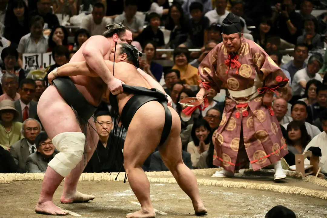 Deux lutteurs de sumo professionnels en plein combat dans le dohyō, sous le regard attentif de l'arbitre traditionnel, lors d'un tournoi au Japon.